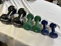 Photo showing all six dumbbells in a row on a white table: two black 10 lb, two green 5 lb, and two blue 3 lb dumbbells.
