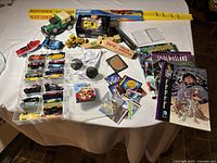 Wide view showing all items on a table including toy cars, train whistles, comics, sunglasses, harmonica, cards, and small collectibles