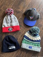 Four fan headgear items displayed on wooden floor: Toronto Raptors knit hat, Toronto Blue Jays baseball cap, Seattle Seahawks knit beanie, Vancouver Canucks knit hat with pom-pom.