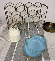 Photo showing all 5 items on a striped cloth: geometric brass wine rack, white watering can, iridescent shot glass, heavy brass jewelry holder, and blue Fire King dish