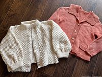Overview of both hand-knit cardigans showing cream knit cardigan with zipped closure and coral button closure cardigan.
