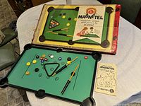 Full view of complete Magnetel magnetic pool skill game set with balls, cues, triangle rack, magnetic stick, new condition magnetic board, box and instruction booklet on table.