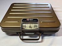 Top front view of Pelouze parcel post scale, showing ridged metal lid and clear weight dial window with branding.