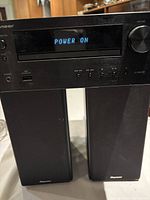 Front view of Pioneer X-HM20 stereo receiver powered on showing digital display with 'POWER ON' message. Two black speakers positioned beside it.