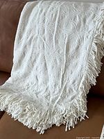 Image of the vintage white or cream bedspread draped over couch showing detailed floral textured pattern and long tassels on edge.