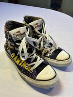 Pair of Converse Chuck Taylor All Star high-top sneakers displayed on a white cloth with colorful Simpsons Batman graphics and white laces.