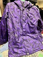 Front view of purple Volcom ski jacket with geometric patterns and hood, laid flat showing zipper and pockets.