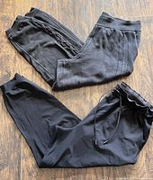 Two pairs of black Lululemon joggers laid flat on a wood floor, showing the front and side views. One pair has braided side detailing.