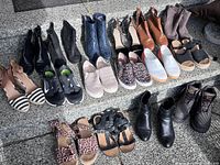 Wide view of 16 pairs of women's shoes in various styles including running shoes, booties, heels, slip-ons, and sandals arranged on a textured floor.