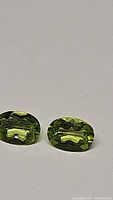 Pair of natural apple green peridot gemstones, oval cut, displayed on a white surface with light reflections showing facet cuts and clarity.