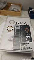 Photo of the ring alongside GRA certification paper and card showing moissanite certification details including size 6.5mm, 1.0 ct weight, D color.