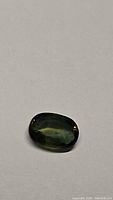 Photo shows an oval cut natural green sapphire gemstone on a plain background. The stone displays a deep, rich green hue with visible facets and transparency.