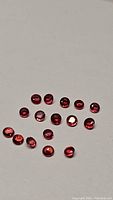 Photo showing 15 small round faceted red rubies with orange undertones on a white background
