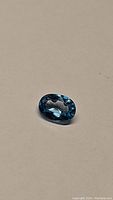 Close-up image of a single oval cut natural blue zircon gemstone placed on a plain surface, showing vivid lagoon blue color and faceted surface.