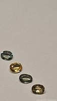 Four natural sapphire gemstones arranged in a row on a plain light background, clearly showing multi hues of green and yellow, with oval cuts and visible facets.