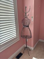 Front view of the black metal wire dress form coat rack placed near a window with white shutters
