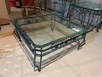 Square coffee table with black metal base featuring decorative scrolling and curved legs, topped with clear beveled glass.