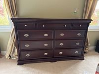 Front view of dark wood dresser showing nine drawers and metal knobs/pulls, placed against a wall with windows and curtains.