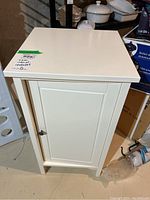 Front view of white IKEA cabinet with one door and metal knob placed on basement floor.