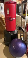 Photo showing full view of red Wavemaster punching bag on black water-fillable base with adjacent purple exercise ball and part of weighted hula hoop visible in background.