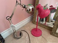 Two desk reading lamps side by side on carpeted floor near pink wall and mirror. One metallic silver with spherical head, one bright pink with two adjustable heads.