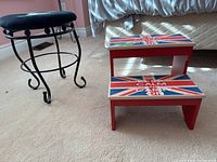 Two stools positioned on carpet. One is a red wooden step stool with British flag and text on steps. The other is a black cushioned seat stool with metal curved legs.
