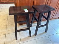 Two dark wood veneer stools standing on tiled floor by wood cabinetry. Shows overall stools design and condition.