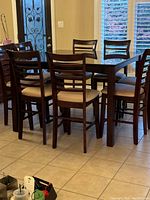 Side angle of rectangular dark wood counter height table with eight matching chairs arranged around it in a tiled dining area near windows.