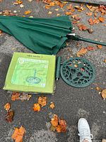 Folded dark green garden umbrella with a metal pole and a disassembled ornate metal stand placed on pavement with autumn leaves.