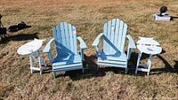 Set of two light blue foldable Adirondack chairs with two small white fish shaped wooden side tables on grassy ground.