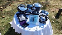 Full view of all cookware items displayed on a white table outside, showing multiple pots with glass lids, grill pan, and lid holder.