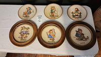 Full view of all seven Hummel plates arranged in two rows, some framed, showing various pastel colored illustrations and visible dates from 1975 to 1991.