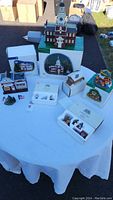 Wide view of all Department 56 Heritage Village porcelain collectibles on a table outdoors, including buildings, boxes, and Amish family figurines in original packaging.