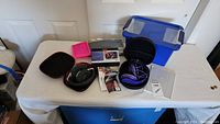 Overview of all items on table: Beats headphones in case, pink magnetic organizer, RCA 3.5" LED TV box, purple headphones in case, blue plastic bin.