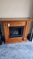 Full view of wooden faux fireplace with decorative carved details, black firebox front with gold trim and faux logs inside.