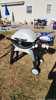 Weber Q gas grill shown assembled outdoors on grass with two wheels, lid closed, side shelves with grill accessories on top including cooking spatula and tongs.
