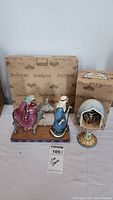 Both Jim Shore collectibles with their original boxes behind them on a white surface, one figurine of Mary and Joseph with donkey and one dome with crucifixion scene on a pedestal