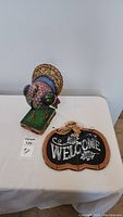 Photo showing Jim Shore colorful turkey figurine and pumpkin welcome sign side by side on a table against a white wall.