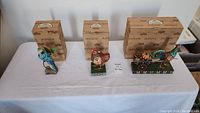 Three boxed Jim Shore figurines displayed on a table: blue parrot, red rooster, and decorative pumpkin cart.