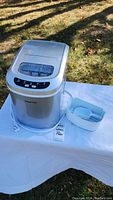 Silver Magic Chef portable ice maker displayed outdoors on white cloth with ice bucket and scoop.