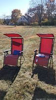 Two red rocking folding chairs with black metal frames standing outdoors on grass with adjustable sun visors open above the seat backs. One chair includes a small blue item on the seat.