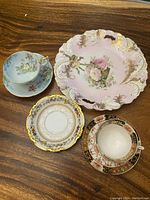 Four porcelain items on wood table: pink floral dish, blue cup and saucer, yellow saucer, brown cup