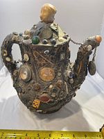 Front and side view showing porcelain doll head finial on lid, collage of buttons, coins and marbles