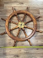 Photo of antique wooden ship's steering wheel with brass hub lying flat on a wooden floor. A measuring tape indicates the diameter is about 32 inches.