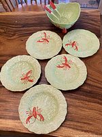 Five pale green textured plates each handpainted with bright red lobster motif, and a matching serving bowl with three red lobster claw utensils inside.