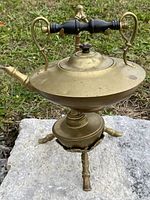 Side view of the brass teapot on the brass tripod stand with wooden handle visible and small burner underneath.