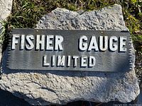 Photo of rectangular cast aluminum sign resting on stone slab, showing raised white lettering 'FISHER GAUGE LIMITED' on textured dark gray background.