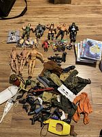 Wide view of vintage toys including multiple G.I. Joe figures, WWF wrestling figurines, clothing, footwear and helmets, alongside stacked Wii video games.
