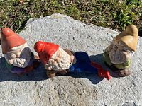 Three vintage garden gnomes positioned on a stone surface outdoors. Each gnome is uniquely colored and posed, showing detail and condition.