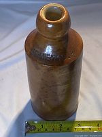 Front angle view of the brown stoneware Schweppes ginger beer bottle with visible embossed branding and measuring tape showing height.